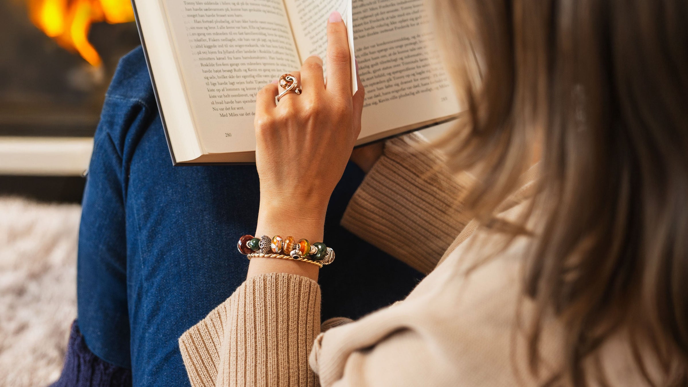 Person, die ein Buch vor einem warmen, gemütlichen Hintergrund liest und dabei die Trollbeads Teestundenzeit Kollektion in Form eines Armbands und eines Rings mit vergoldeter Armspange trägt.