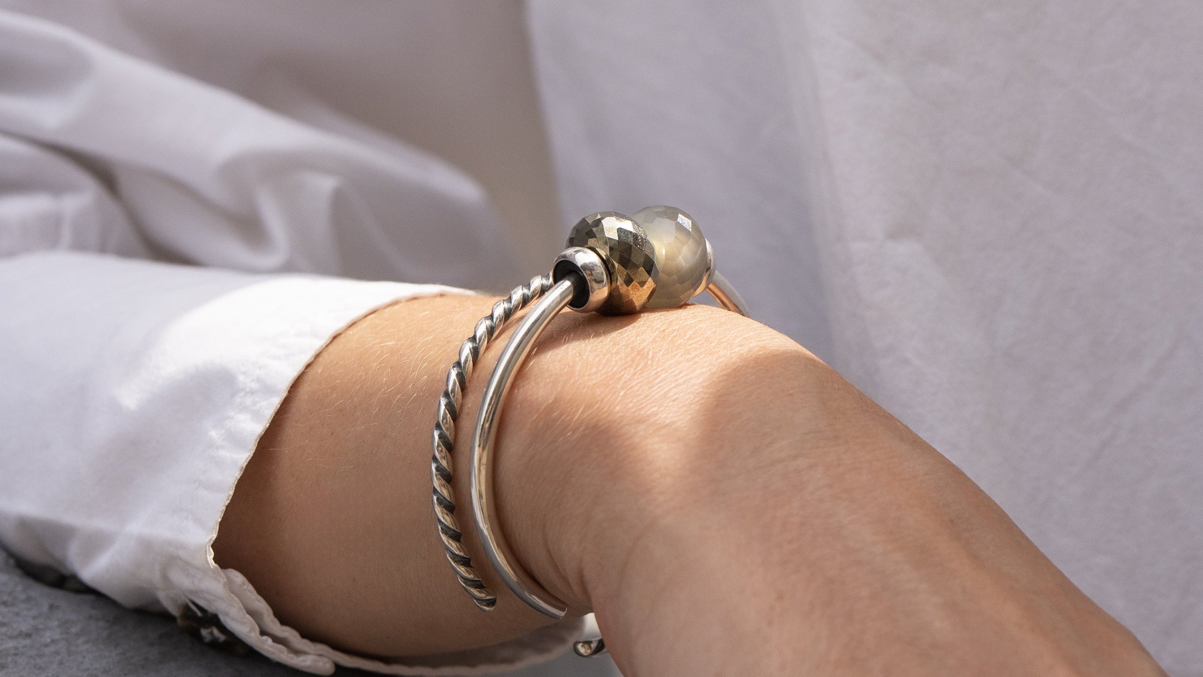 Nahaufnahme eines Handgelenks, an dem zwei silberne Armbänder mit grauen und goldenen Beads auf weißem Hintergrund zu sehen sind